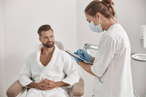 Young administrator in protective mask and gloves fills form on clipborad examining male patient in contemporary cosmetologycal clinic side view